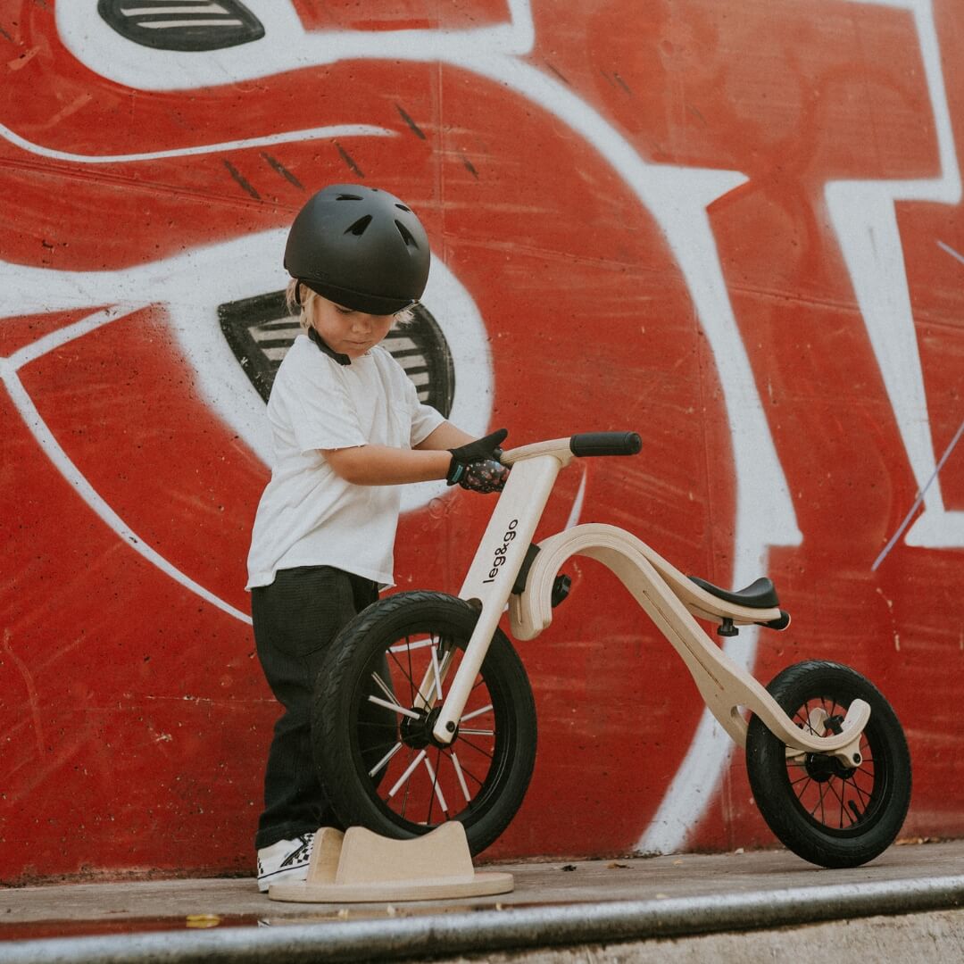 Child wearing a black helmet standing next to a balance bike against a red graffiti wall