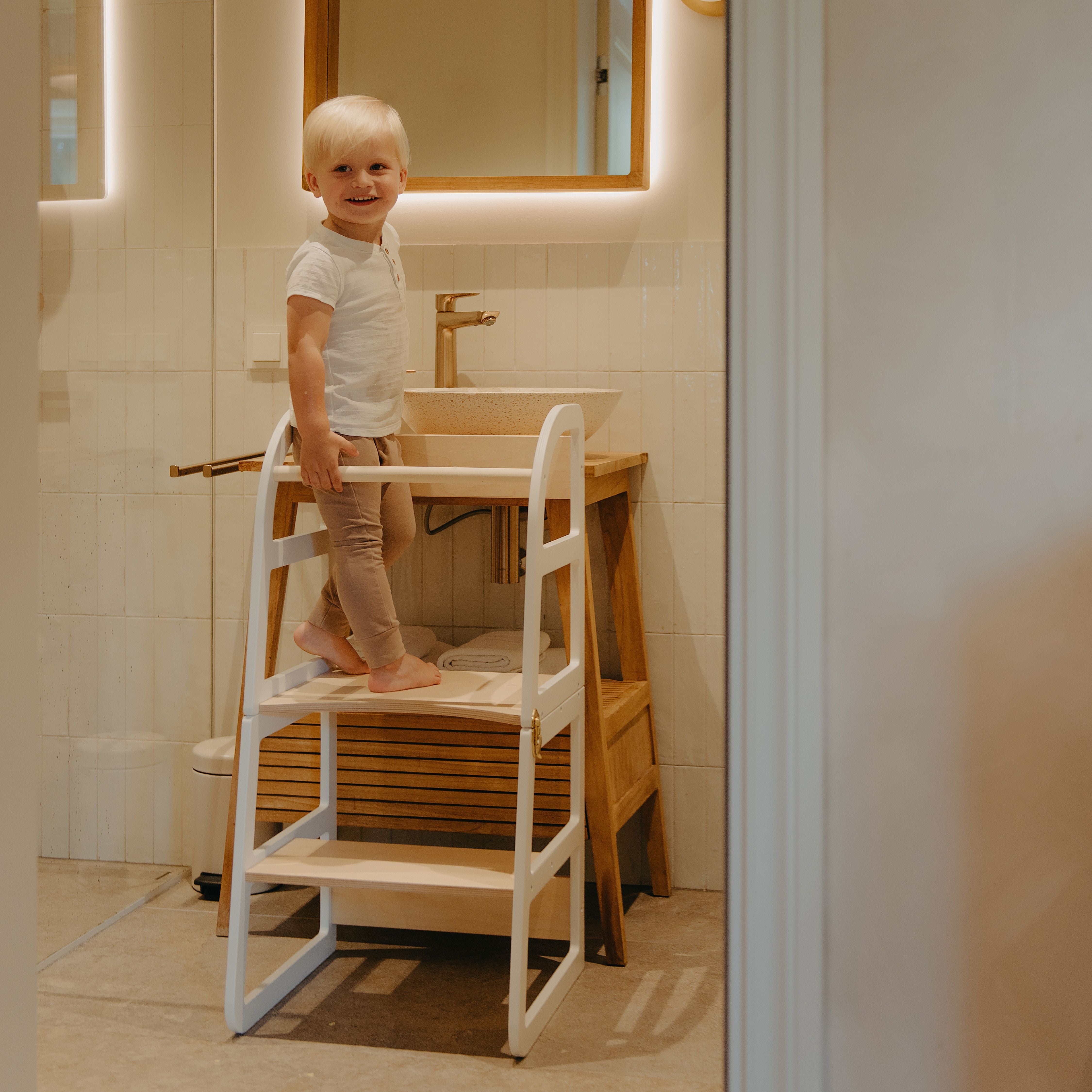 Child standing on a white transformable kitchen tower to independently reach the bathroom sink