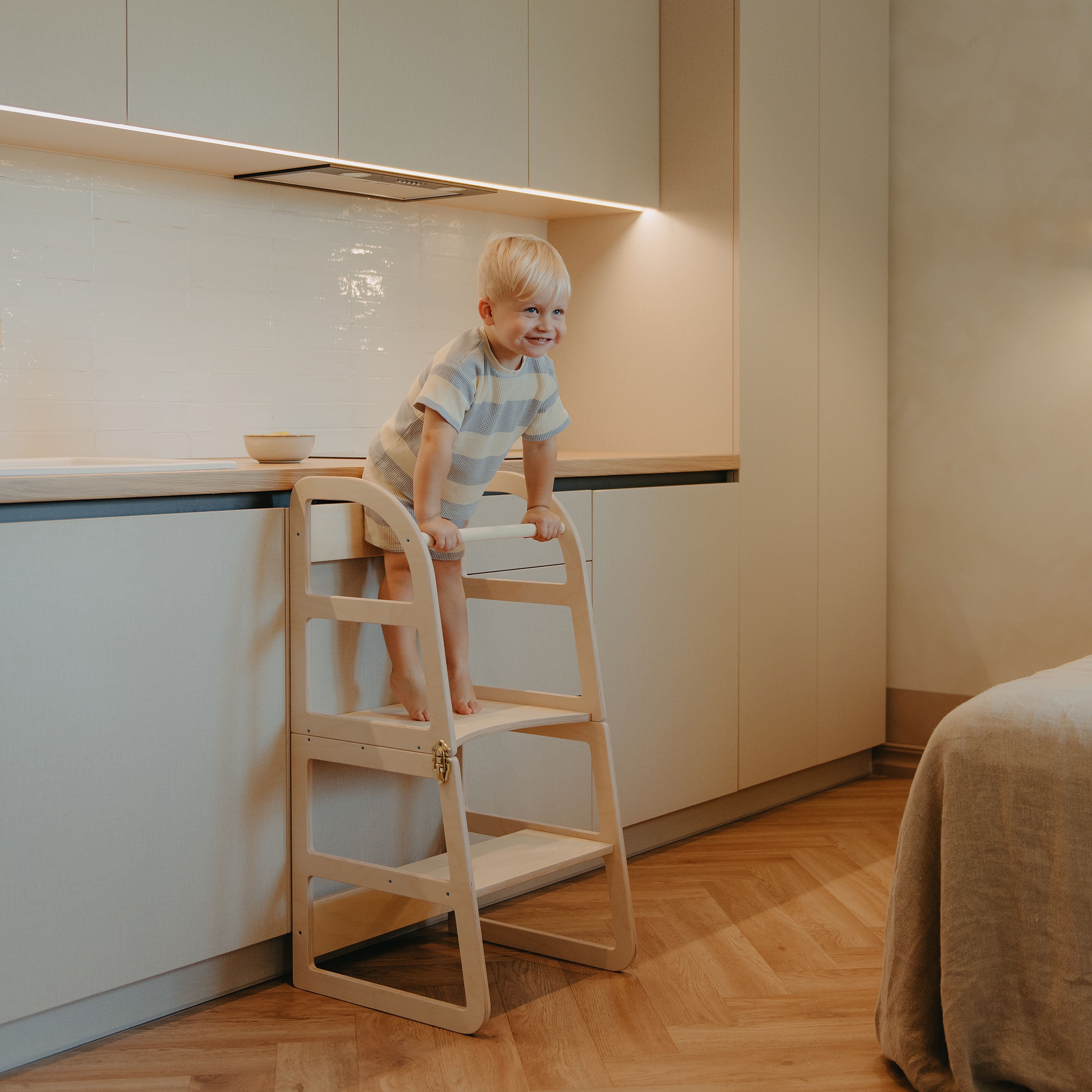 Toddler standing safely on a natural transformable kitchen tower to access the kitchen counter for independent play