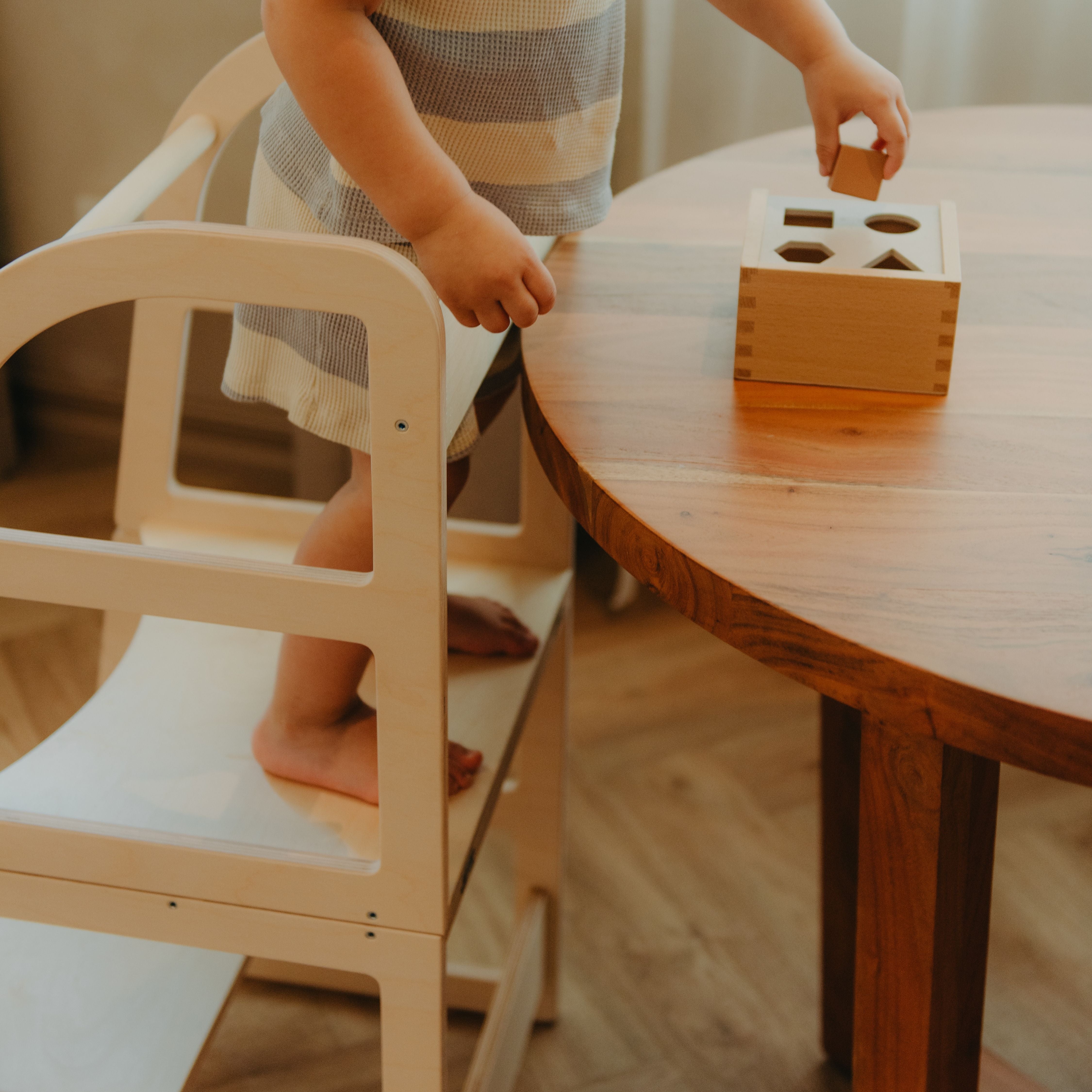 Child using a transformable kitchen tower to reach the table and play with a shape sorting toy for early learning.