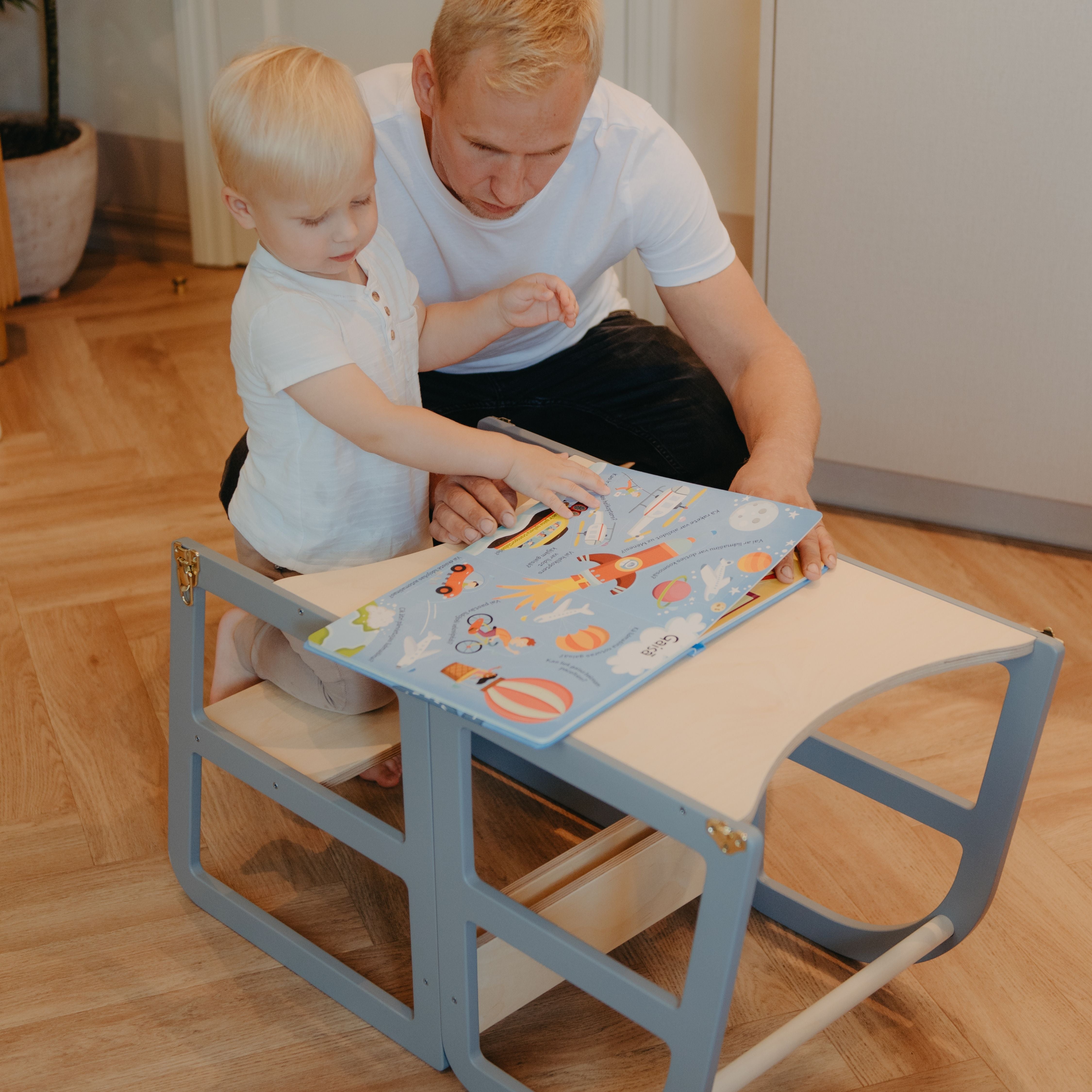 Toddler using a transformable kitchen tower converted into a table for learning and reading with a parent