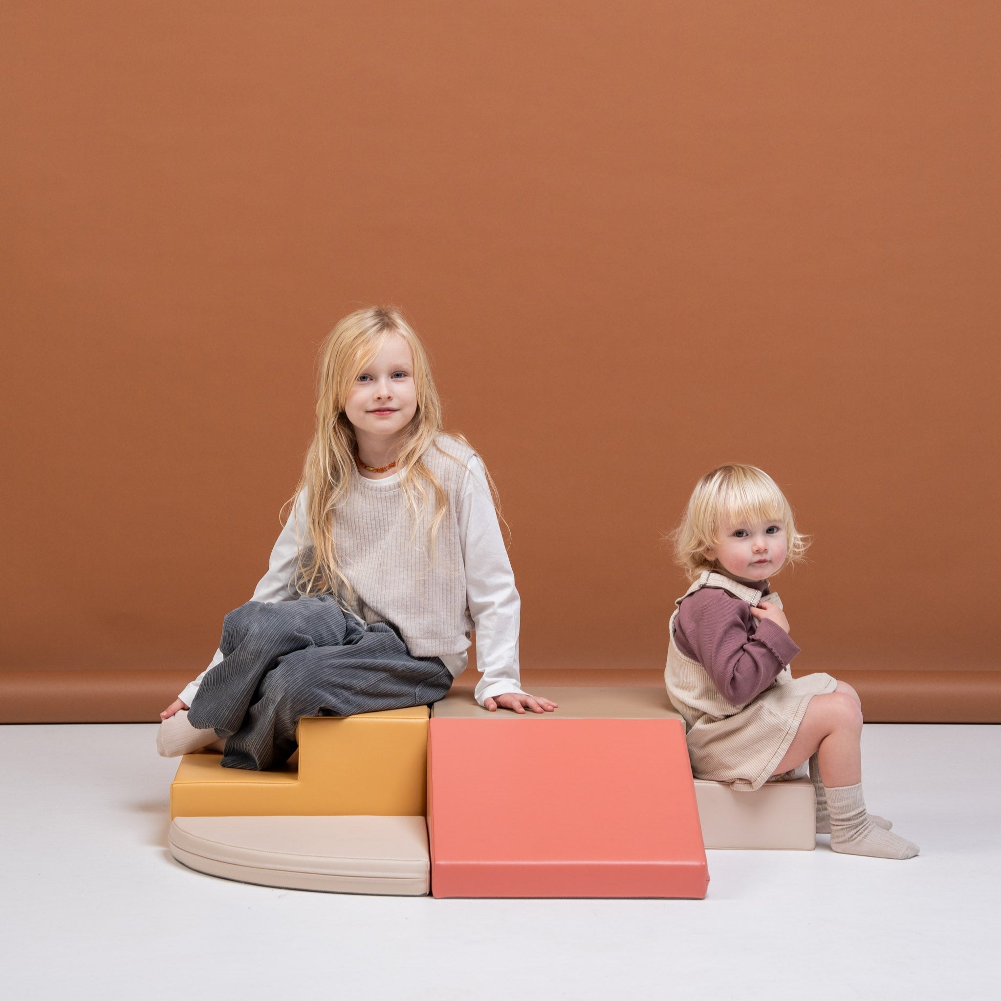 Two children sitting on a beige and pink soft play corner climber set, engaging in calm play