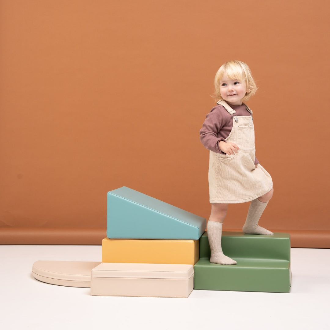 Girl standing on beige and turquoise soft play corner climber blocks, practicing balance and gross motor skills