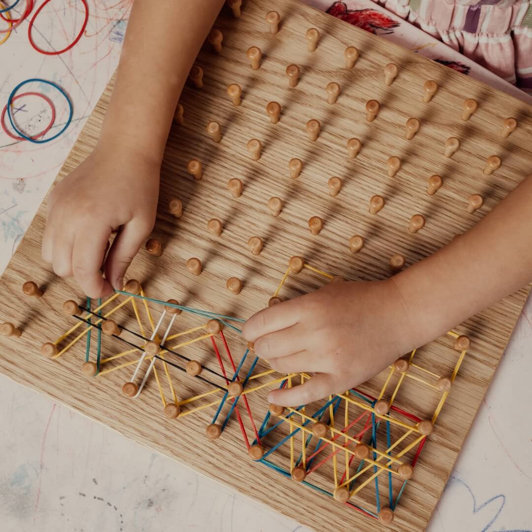 Hands arranging rubber bands on wooden Montessori geoboard – math manipulative for homeschooling