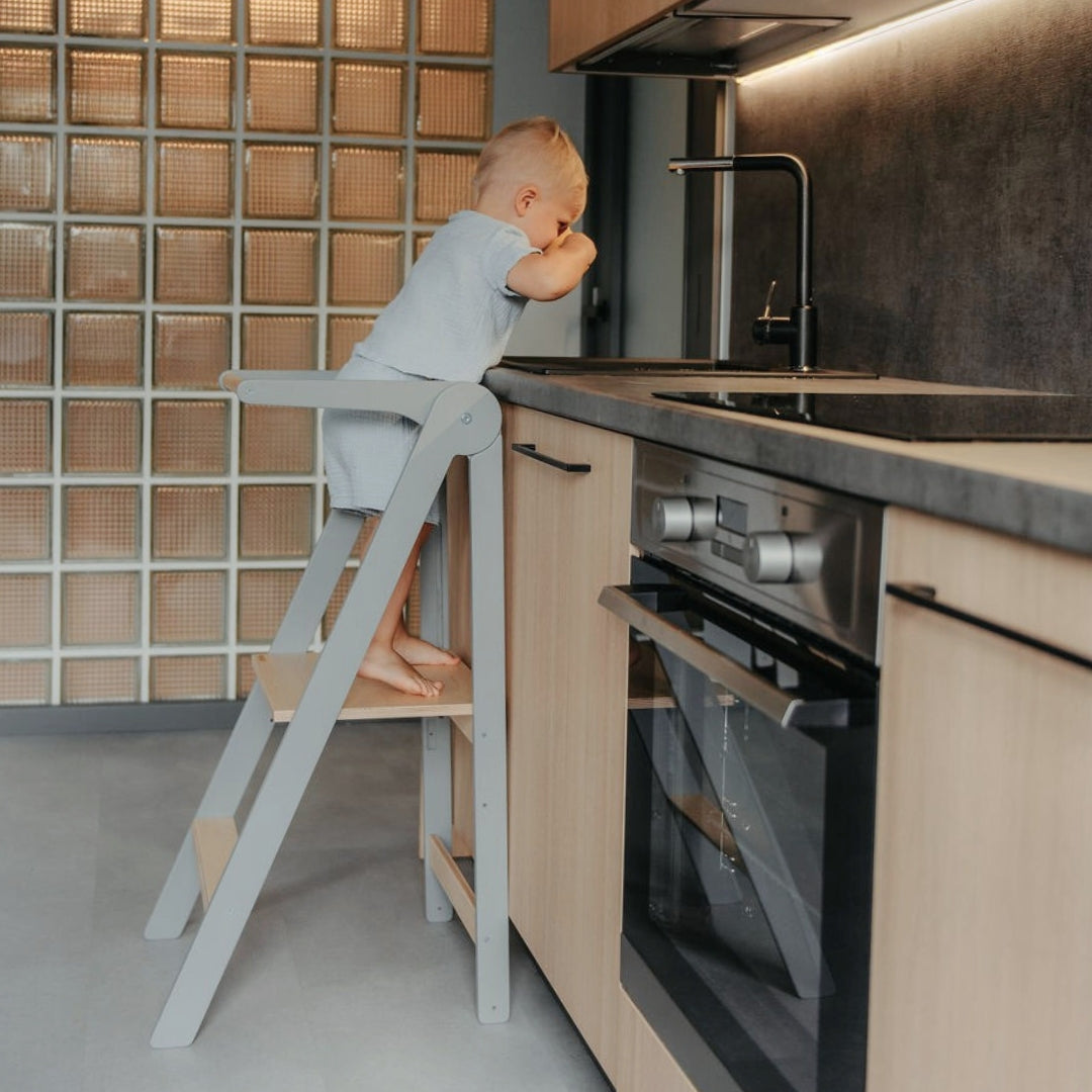 Child standing on a grey kitchen helper tower in a modern kitchen, reaching the water tap safely