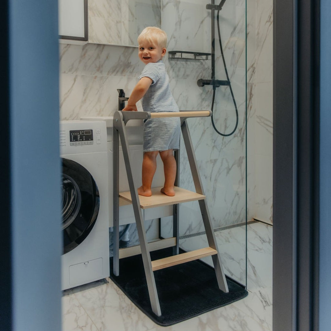 Child standing on a gray helper tower  in a bathroom with marble walls