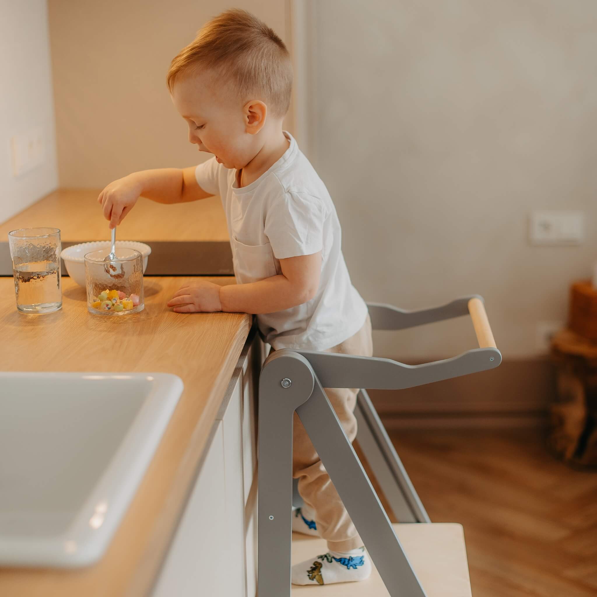 Toddler standing in foldable learning tower at kitchen counter with safety rail