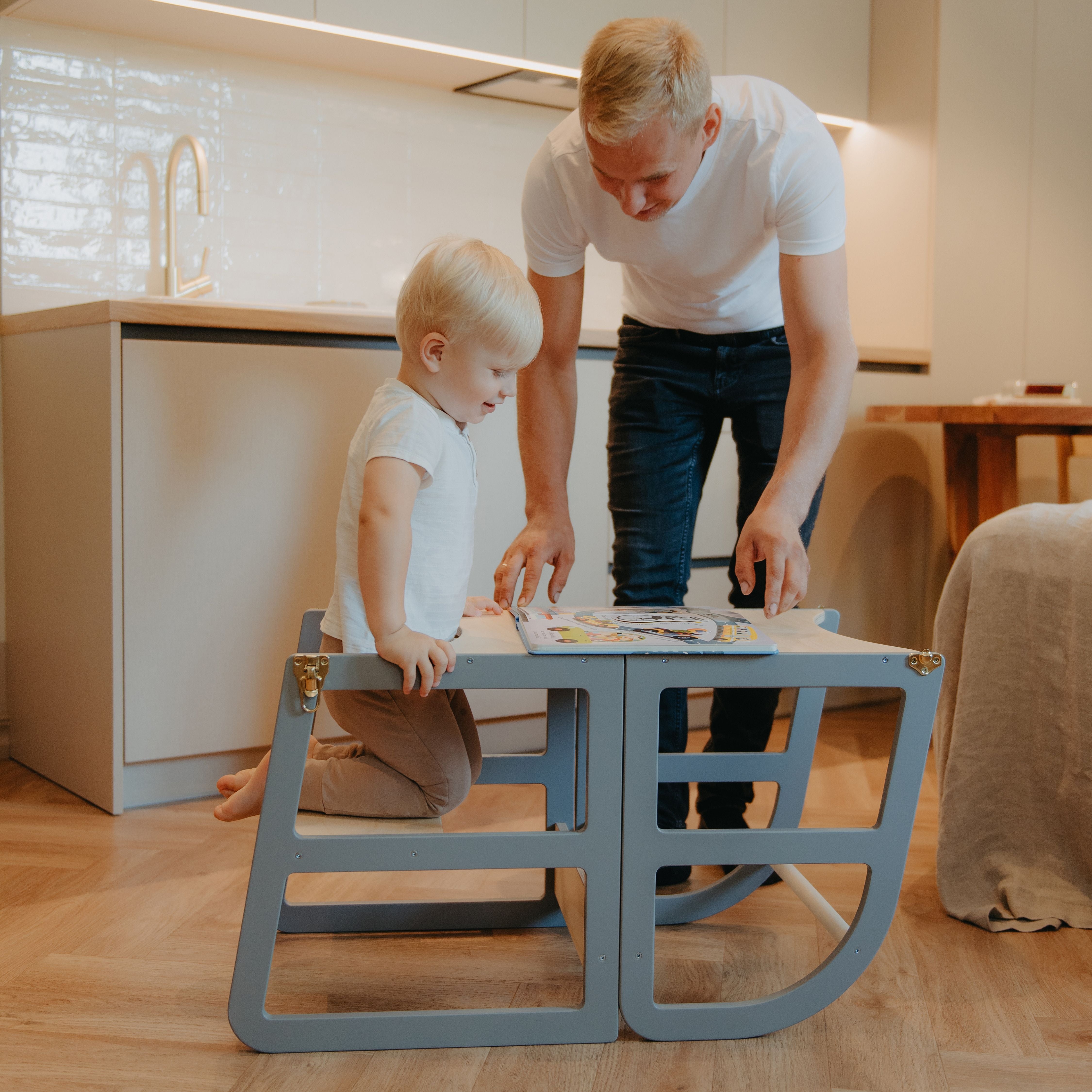 Toddler using a grey transformable kitchen tower as a table while reading with a parent