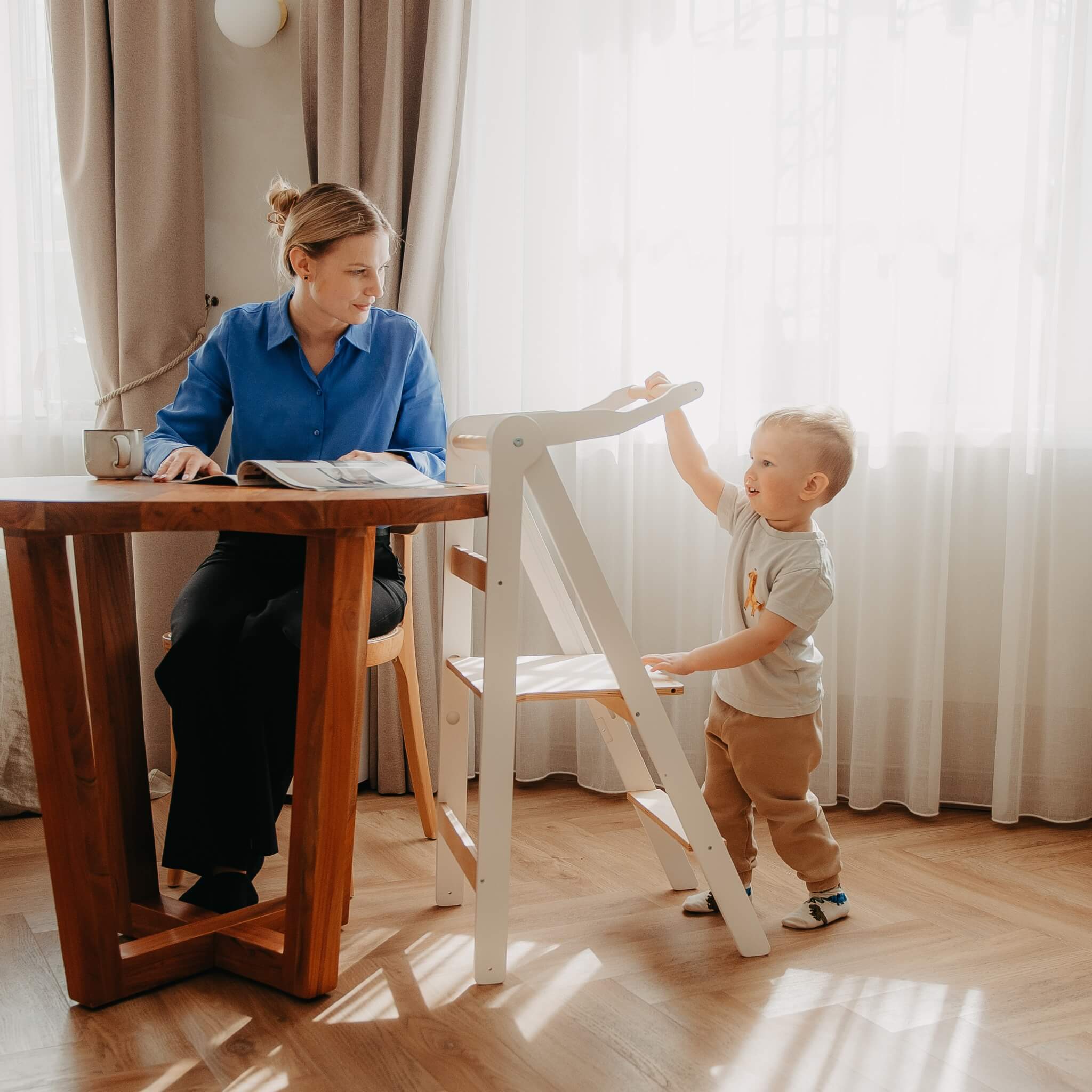Child standing in wooden learning tower interacting with parent in kitchen environment
