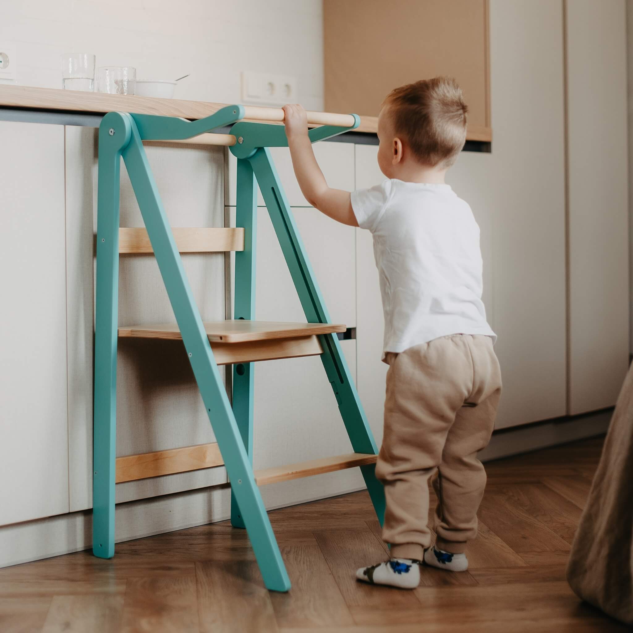 Toddler standing in turquoise wooden learning tower with safety rail
