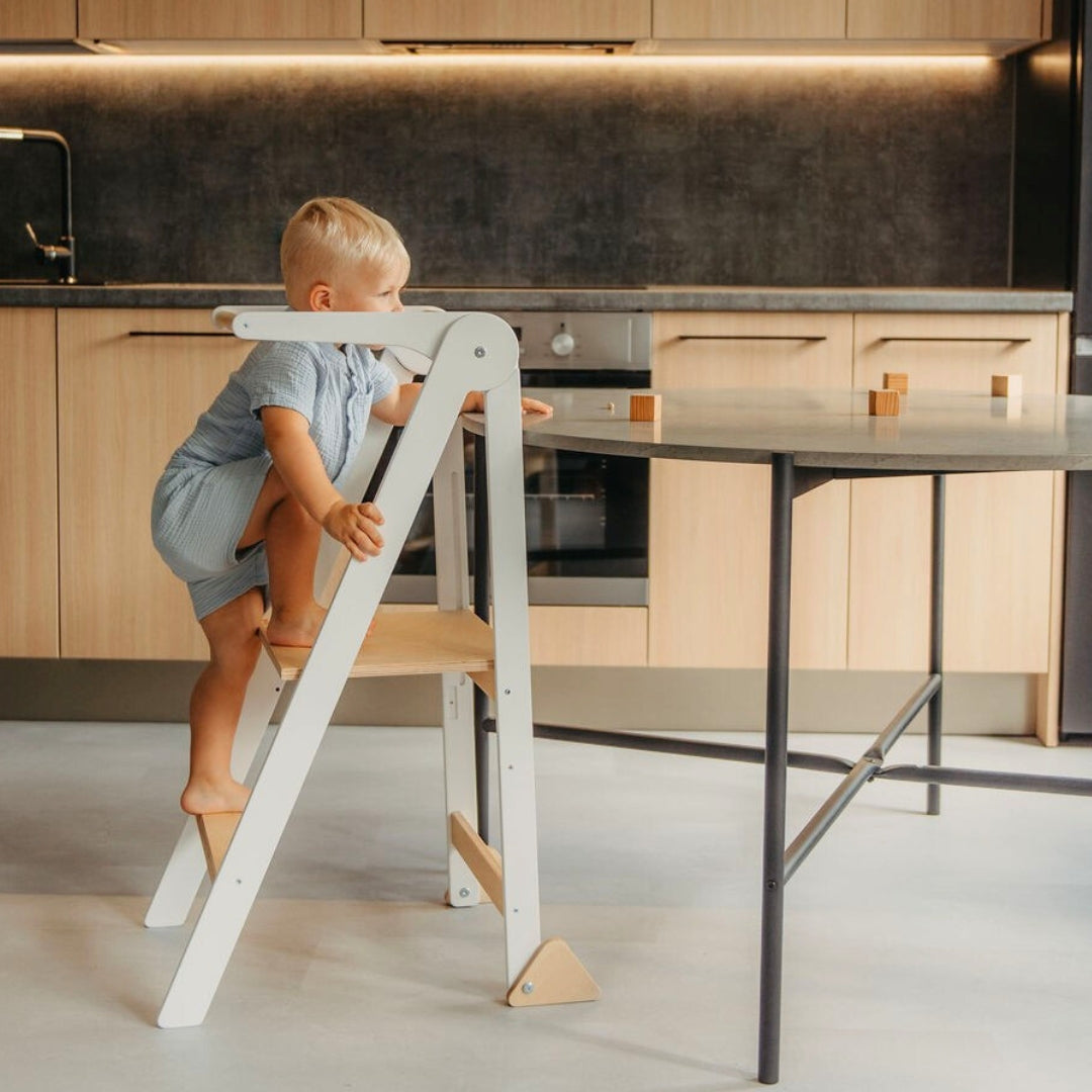 Child climbing on a white kitchen tower to reach a table with wooden play blocks